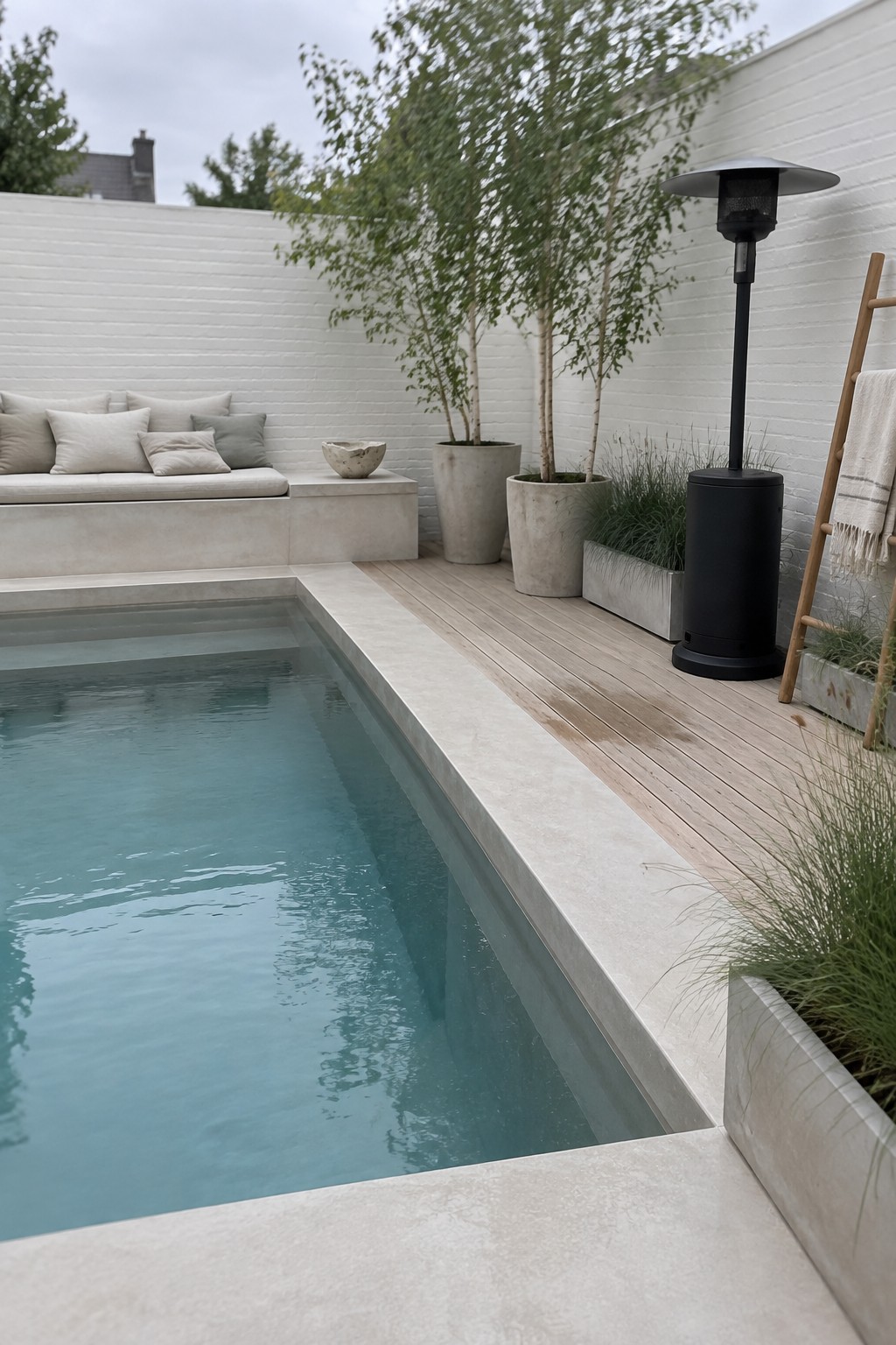 A narrow plunge pool with concrete coping runs beside a wooden deck, with built-in cushioned seating against a white brick wall and several large potted trees nearby.