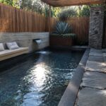 A rectangular plunge pool with dark water sits beside a path of large gray stone pavers, with a concrete bench and small waterfall on the left and a wooden pergola overhead.