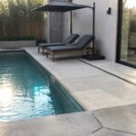 A long narrow plunge pool with blue water next to a concrete patio, two gray lounge chairs, a bamboo screen, and an outdoor shower set into a recessed wall.