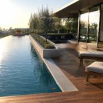 A long infinity pool runs parallel to a wooden deck with two lounge chairs, next to a modern house with large glass doors opening to an outdoor seating area and fire pit.