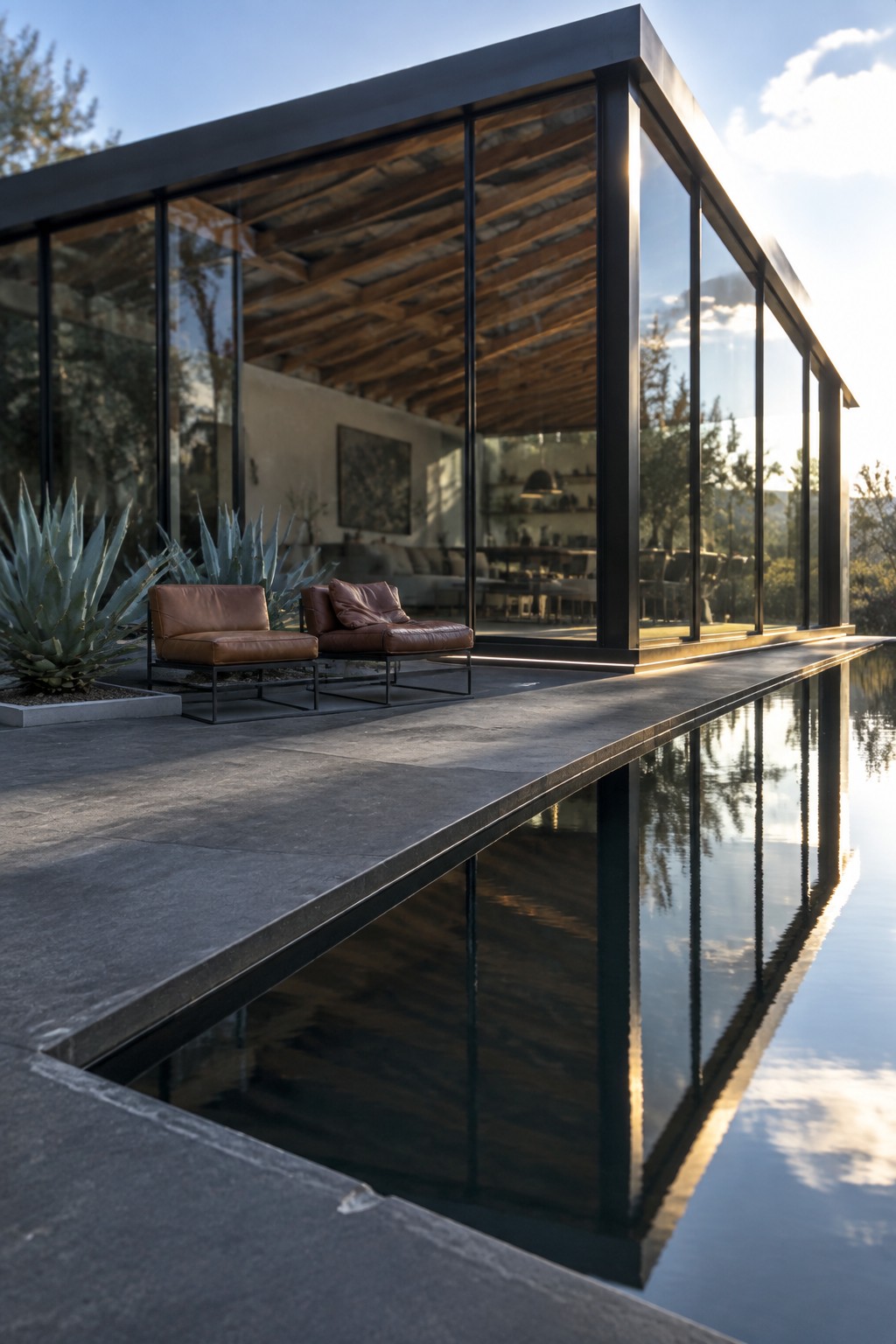 18 Sophisticated Glass House Swimming Pool Designs With Seamless Views A modern glass house with black frames and large windows sits above a long rectangular reflecting pool, with two brown leather lounge chairs on the concrete deck next to agave plants.