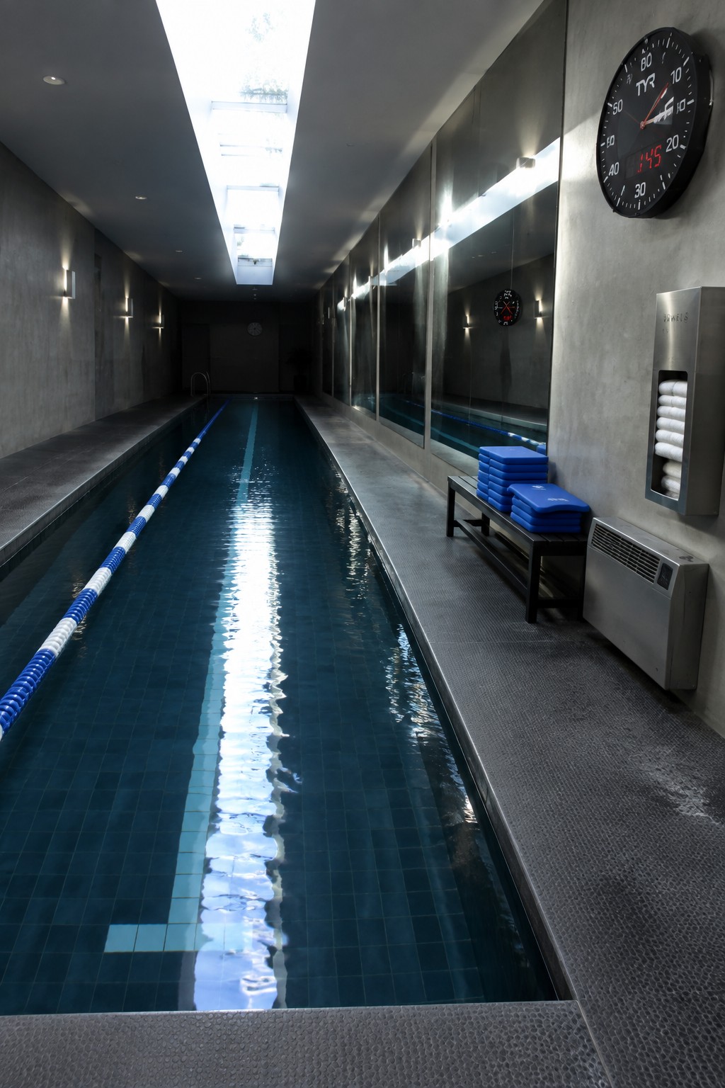 An indoor lap pool with a long rectangular skylight overhead, blue lane lines on the pool floor, and mirrors along one wall.