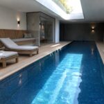 Modern indoor lap pool with blue mosaic tiles, sunlit water, and wooden lounge chairs