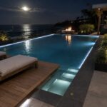 An infinity pool at night with LED strip lighting along the water edge, a fire pit on the left, lounge chairs on a wooden deck, and an outdoor bar area overlooking the ocean under moonlight.