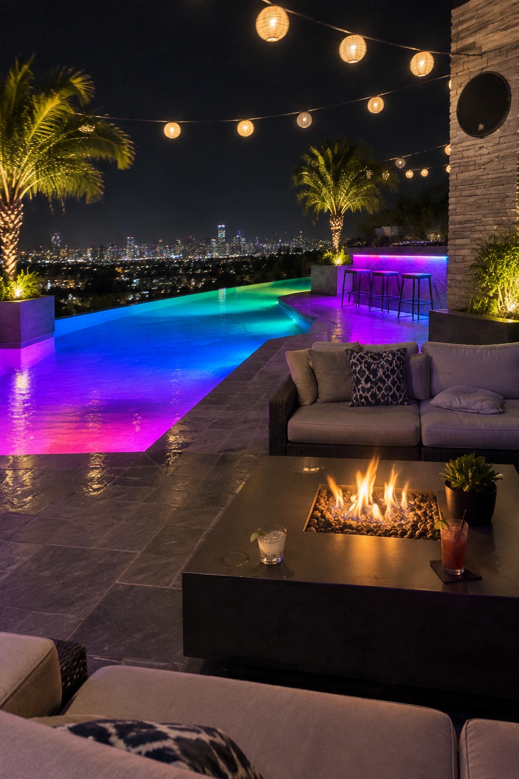 An infinity pool at night lit with blue to purple gradient LED lighting, with a dark fire pit table, gray outdoor sofas, string lights, and palm trees overlooking a city skyline.
