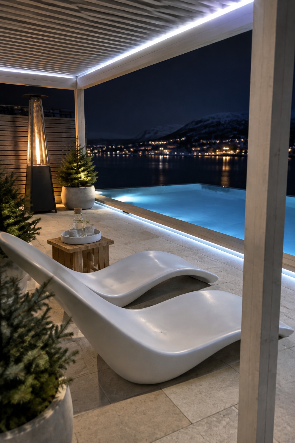 A nighttime view of a modern outdoor patio with two white curved lounge chairs, a glowing infinity pool edged with LED lighting, a tall patio heater, potted trees, and a distant view of water and lights.