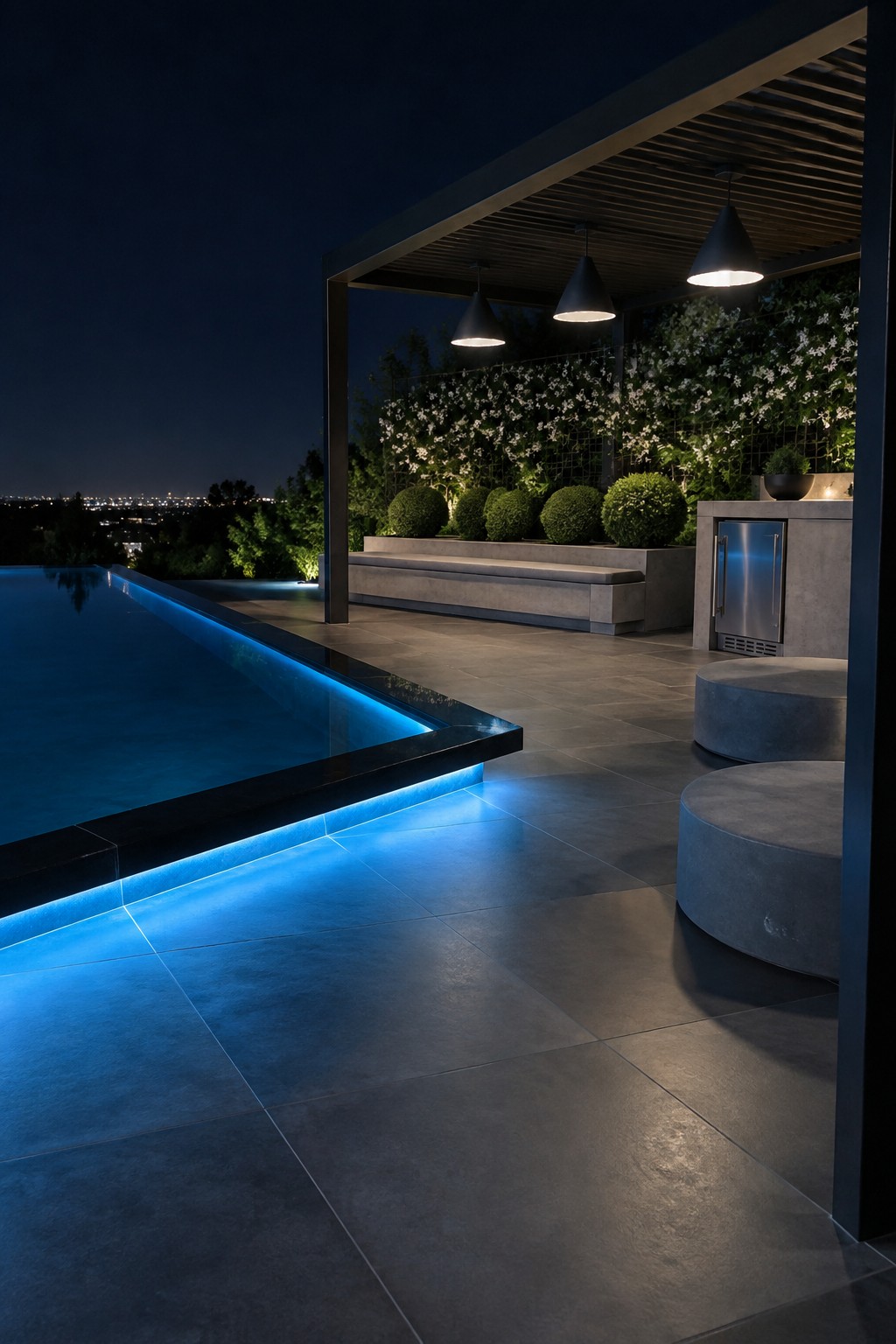 Nighttime view of an infinity pool with blue LED strip lighting along the edge, dark stone tiles, and a covered patio area with round concrete stools.