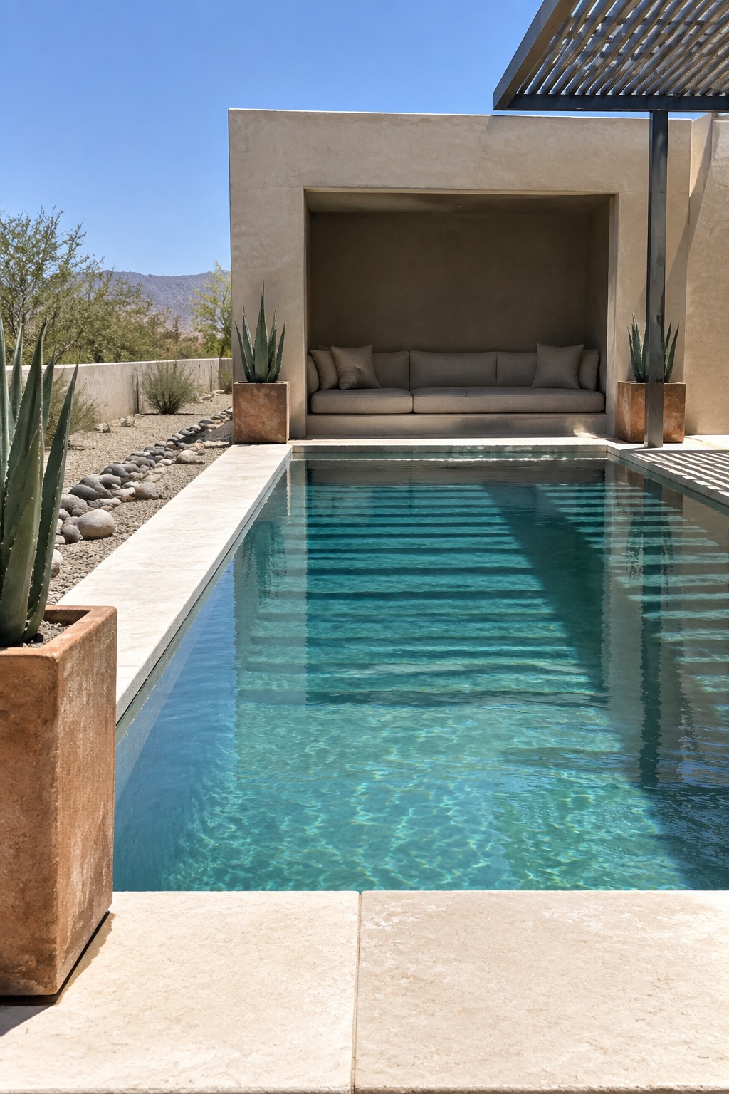 A rectangular modern pool with a recessed seating niche containing beige cushions, flanked by two potted agave plants in terracotta containers and a partial overhead pergola.