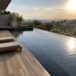 A long rectangular infinity pool with a glass edge sits next to a wide wooden deck holding two light-colored lounge chairs, with a built-in fire pit and distant city and water views in the background.