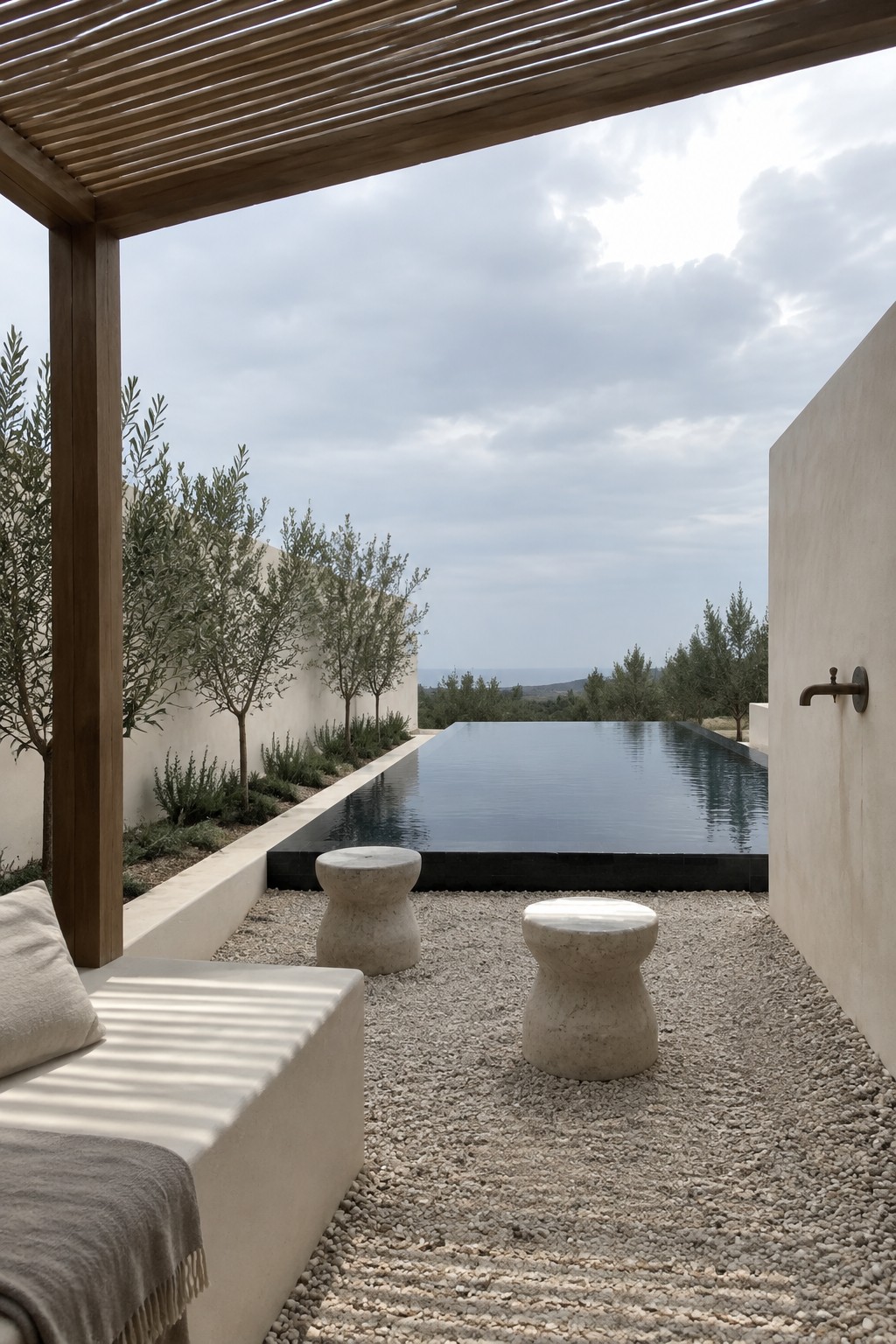 An infinity pool with dark edging sits beside a gravel area holding two light stone stools, a partial daybed under wooden beams, and a wall-mounted faucet, with trees and distant hills in view.