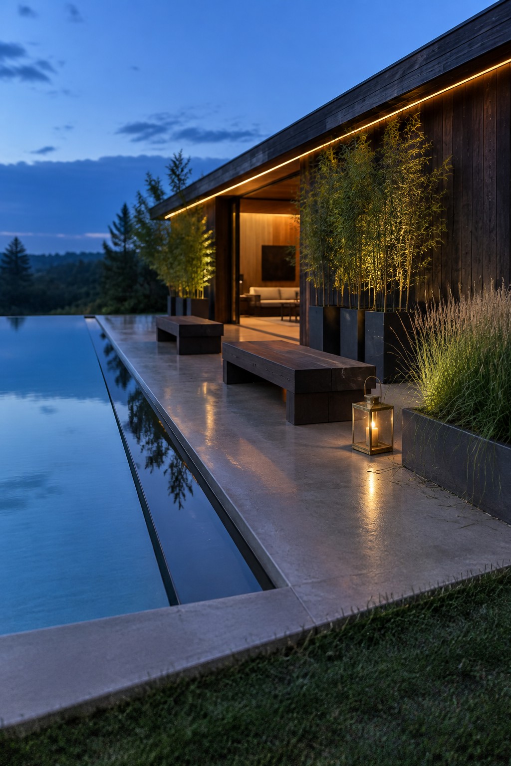 A modern dark wood house next to a long infinity pool at dusk, with wooden benches on the concrete patio and a lit lantern.