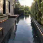 A narrow rectangular lap pool with dark tile lining, a built-in bench with green cushions along the left concrete wall, and a tall vertical garden wall on the right, seen from a dark wood deck at sunset.
