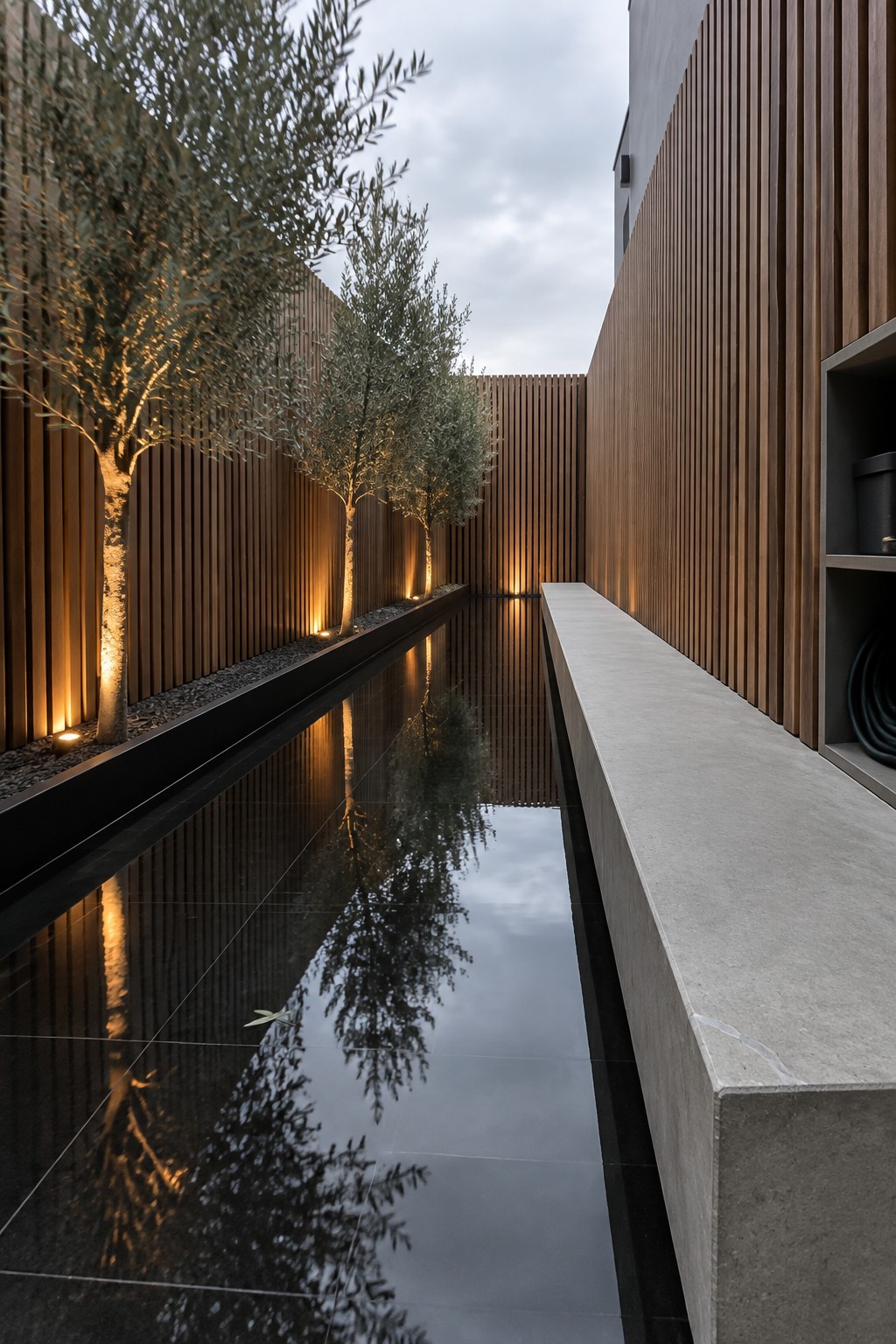 16 Smart Infinity Pool Designs for Narrow Yards A long narrow reflecting pool runs between wooden slat walls with olive trees lit from below and a concrete bench along one edge.