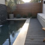 Modern backyard plunge pool with waterfall, wooden deck, concrete bench and plants