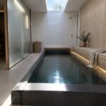 A narrow indoor plunge pool with dark water runs between white tiled walls and a light wood bench, lit by a skylight overhead.