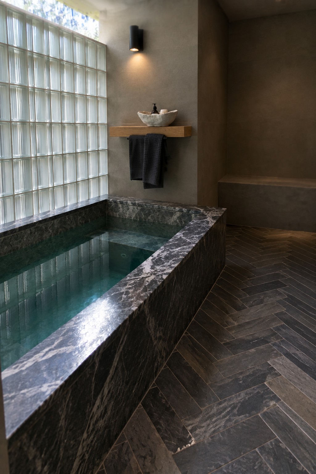 An indoor plunge pool with dark marble coping runs along a glass block wall, with a floating wooden shelf holding a stone vessel sink and a dark towel hanging below it.