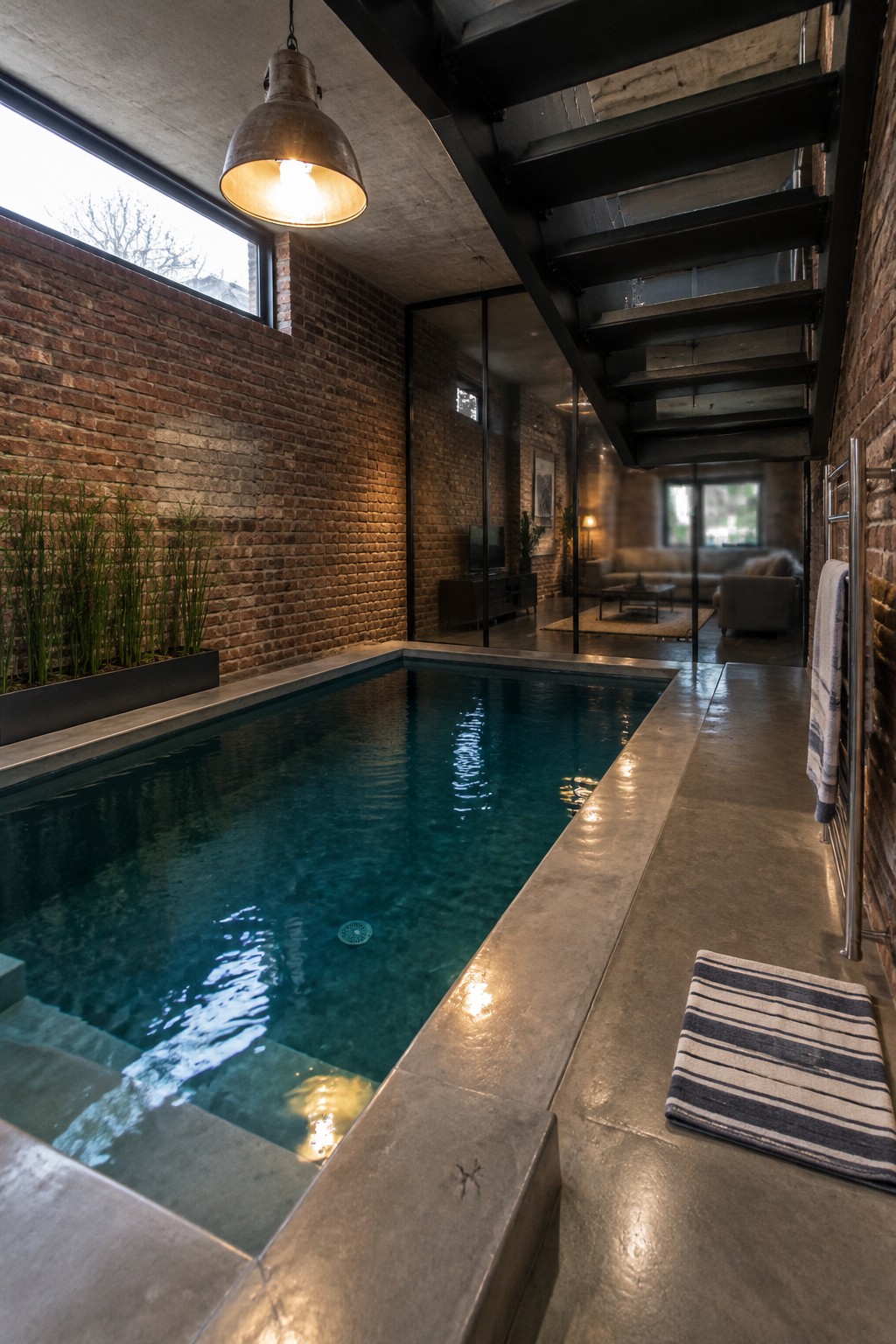 An indoor plunge pool with blue-green water runs along a brick wall beside a concrete walkway under a black metal staircase.