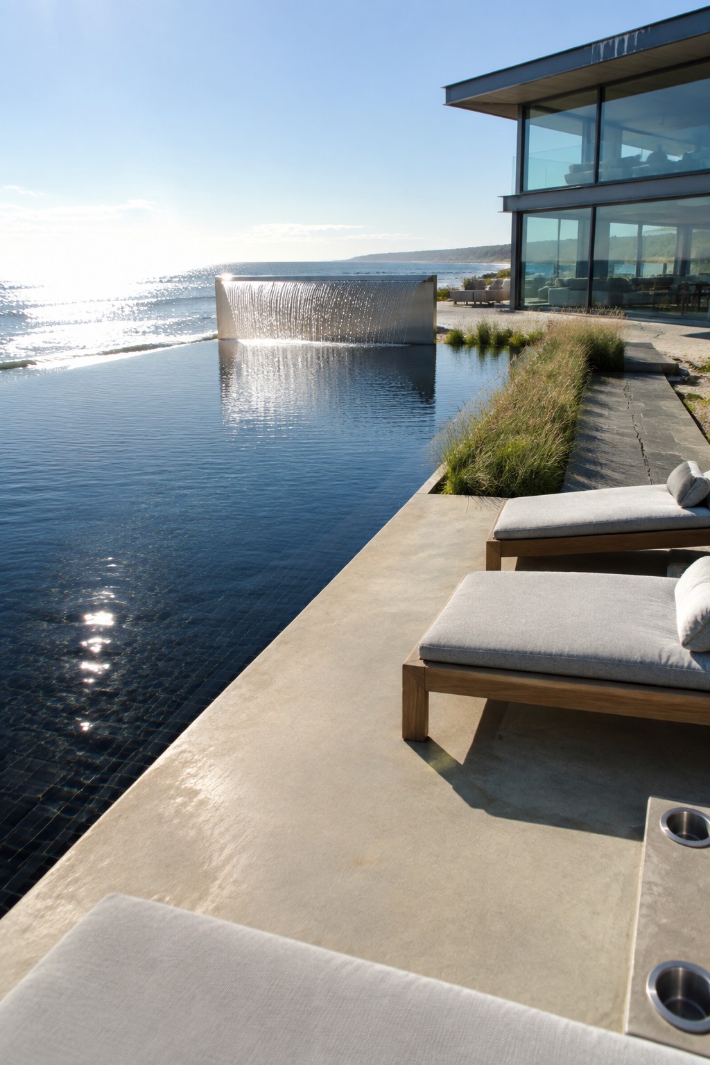 22 Breathtaking Ocean-View Infinity Pools Framed by Coastal Landscapes A long infinity pool with a metal waterfall feature, two wooden lounge chairs on a concrete deck beside the water, and a modern glass house overlooking the ocean.