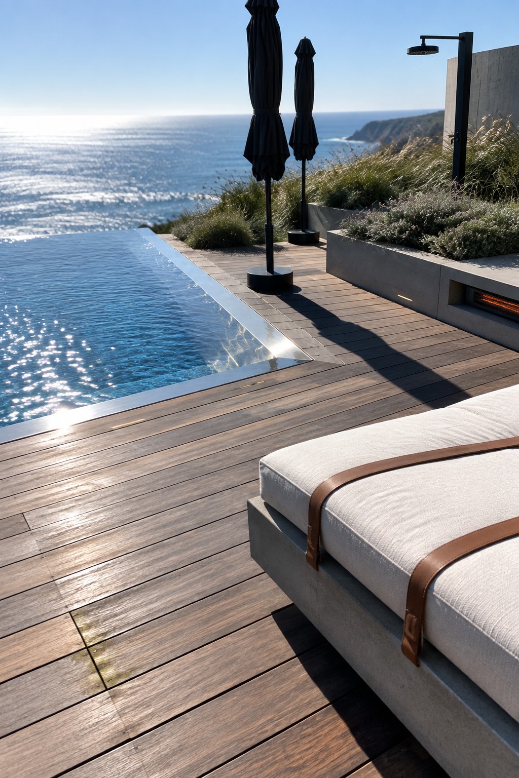 22 Breathtaking Ocean-View Infinity Pools Framed by Coastal Landscapes A dark wood deck next to an infinity pool with a white lounge chair, black umbrellas, and ocean view beyond.