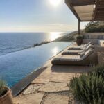 A wooden deck with four lounge chairs in a row beside an infinity pool overlooking the ocean, with stone steps and coastal plants nearby.