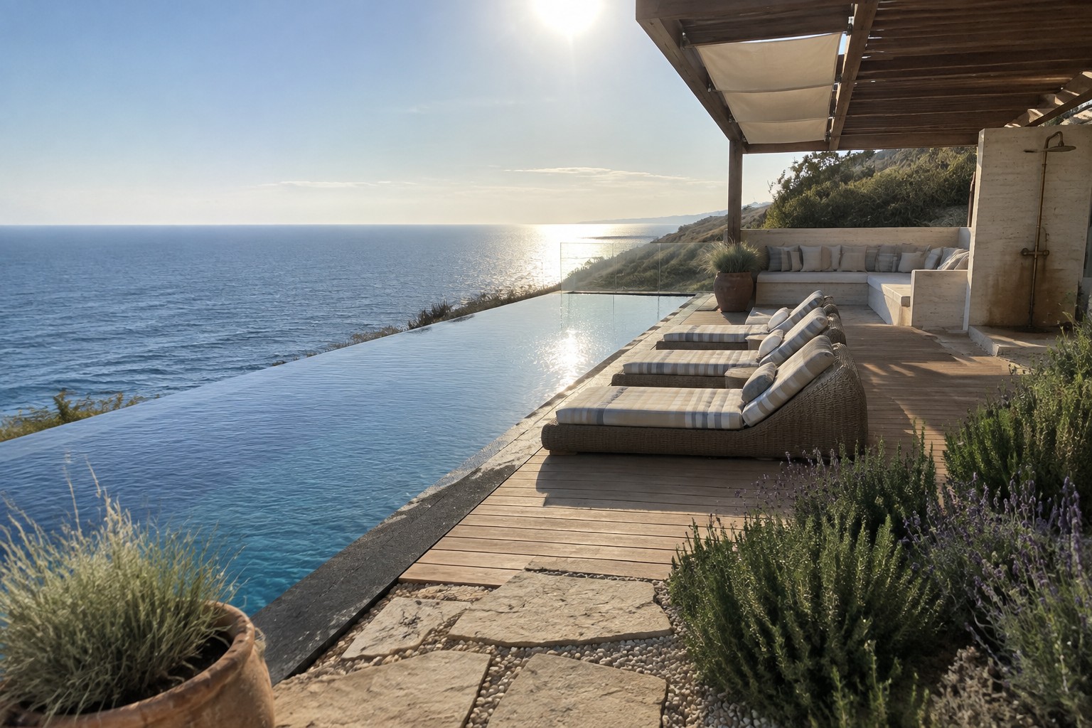 A wooden deck with four lounge chairs in a row beside an infinity pool overlooking the ocean, with stone steps and coastal plants nearby.