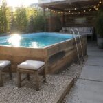 A rectangular plunge pool enclosed by wooden planks with blue tile rim, metal ladder, gravel path, stone pavers, and an outdoor shower on the side.