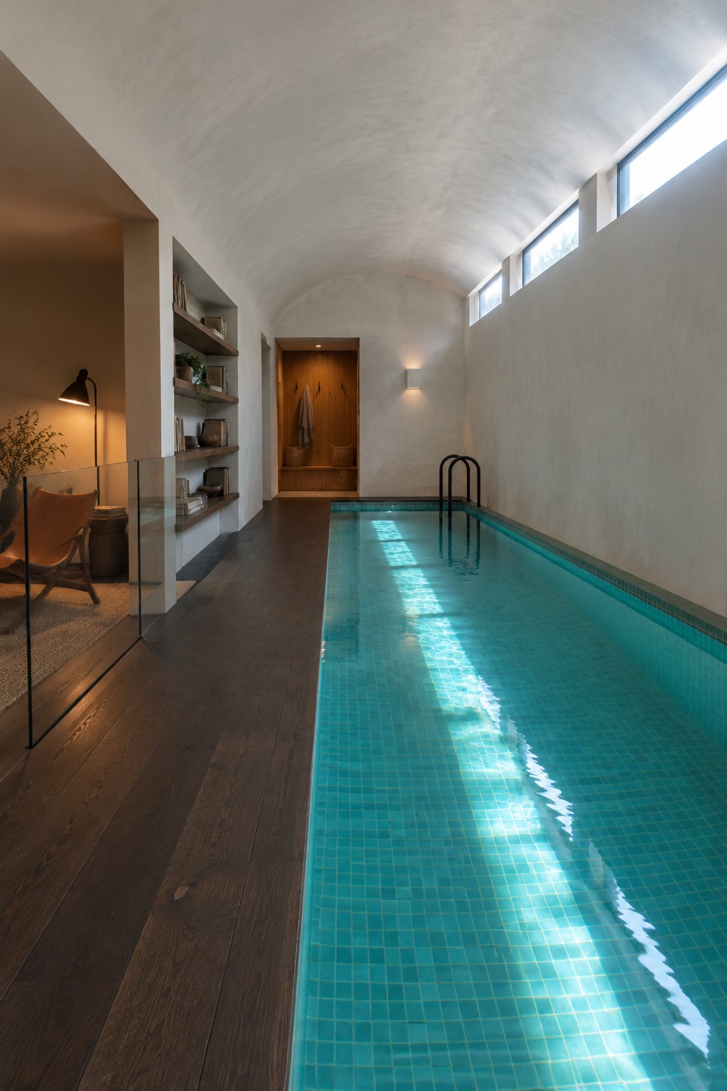 A long narrow indoor lap pool with turquoise mosaic tiles runs the length of a vaulted room beside built-in wooden shelves and a glass partition leading to a seating area.