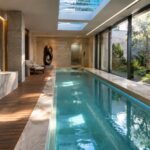 Luxury indoor lap pool with turquoise water, skylight, stone walls, and wooden deck