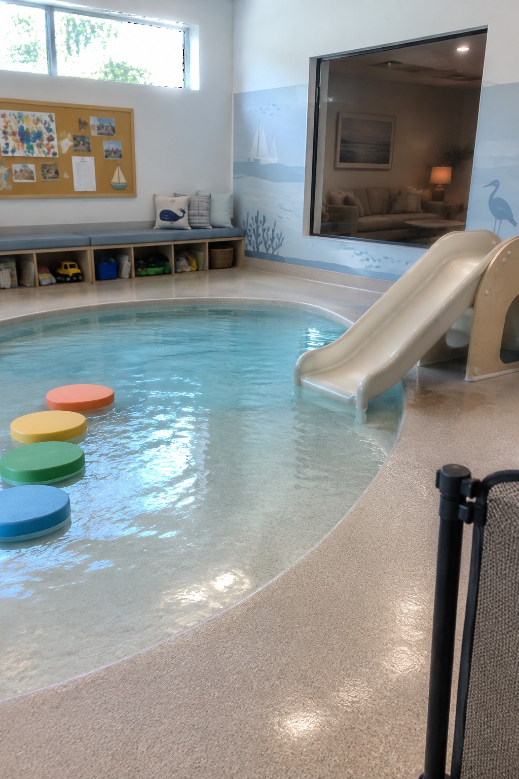 An indoor pool room with a beige waterslide on the right, colorful floating stepping stones in the water, and a large window looking into an adjacent sitting area.