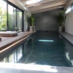An indoor pool with dark stone tile, a light wood raised deck on the left holding two lounge chairs and potted palms, and large glass doors on the right overlooking a garden.