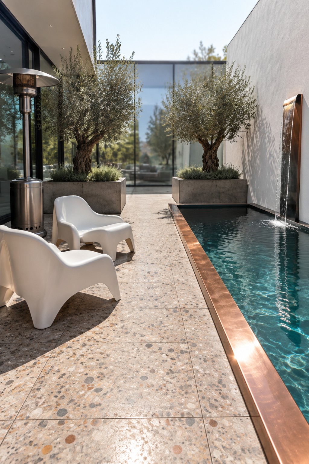 22 Stunning Plunge Pool Ideas for Small Courtyards A narrow rectangular plunge pool with copper edging sits beside a stone-tiled courtyard with two white molded chairs and olive trees in concrete planters.